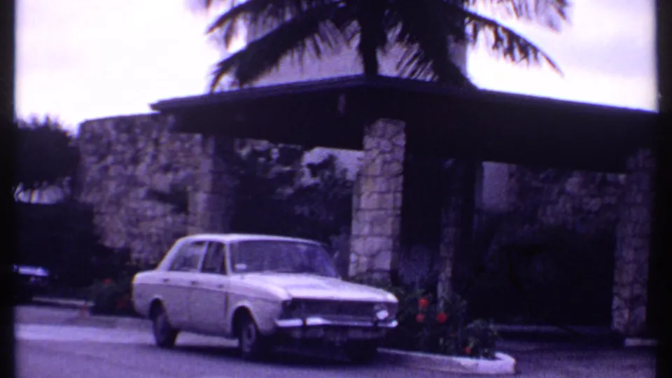 1969 Bahamas home-movie frame showing a white sedan by a resort entrance with stone columns, a palm tree, and planted flowers.