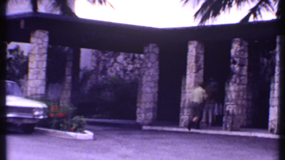 1969 Bahamas frame showing a person near a covered resort entrance, with stone pillars, landscaping, and part of a white car in view.