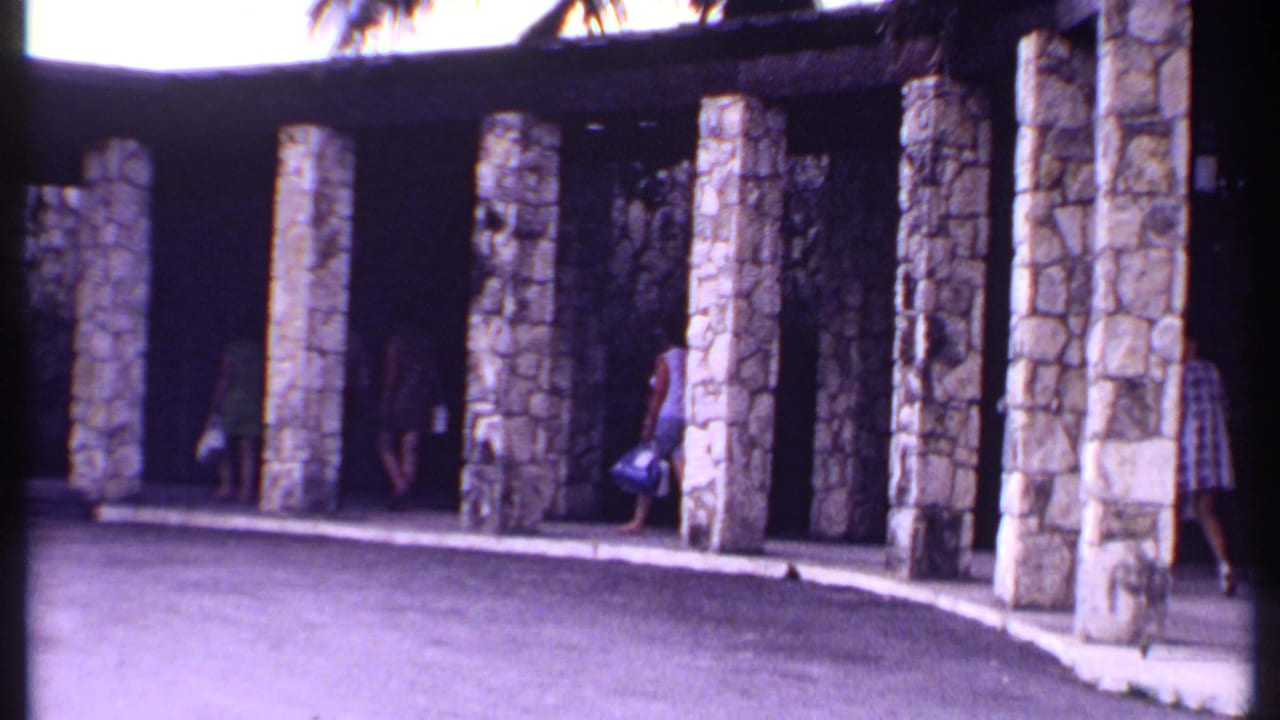 1969 Bahamas home-movie frame showing travelers under a stone-columned resort entrance with luggage and palms above.