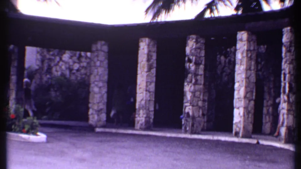 1969 Bahamas resort entrance with stone pillars, shaded walkway, greenery, and partially visible pedestrians.