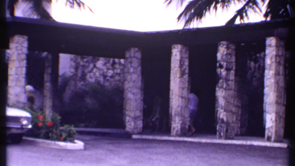 1969 Bahamas home-movie frame with a white car, stone-pillared resort entrance, flowers, and people in shade.
