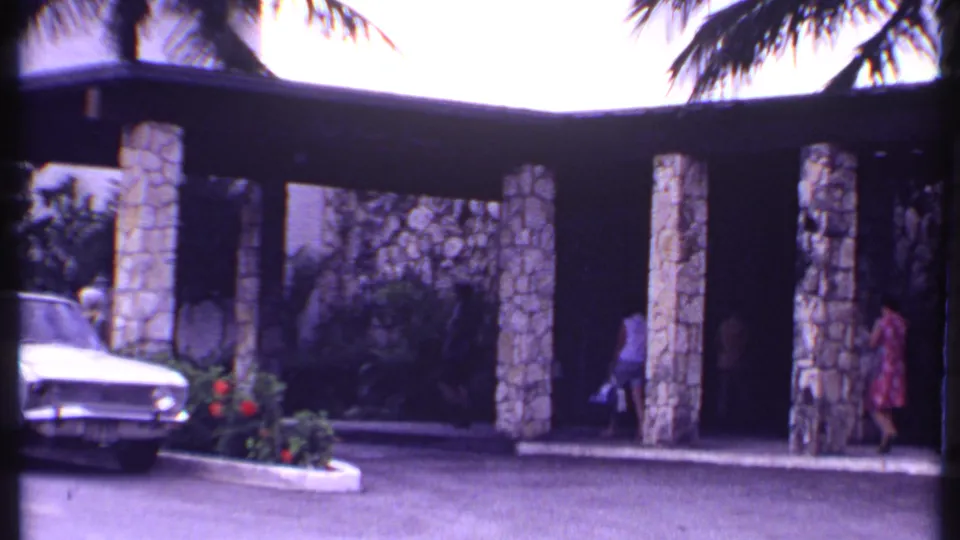 1969 Bahamas resort arrival scene with women walking near luggage, stone columns, palms, and a parked white car.