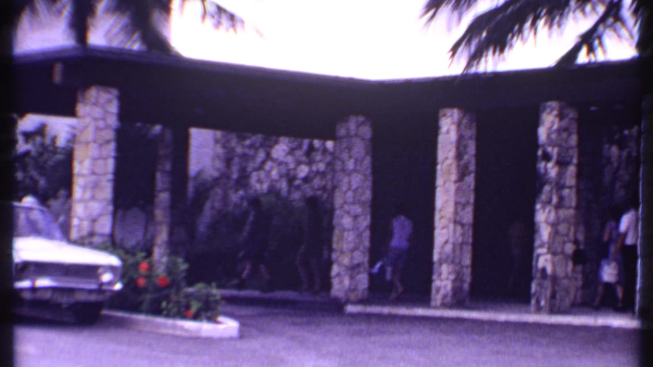 Stone resort entrance with palm trees, a white sedan, and distant pedestrians.