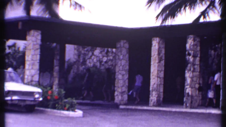 1969 Bahamas 8mm frame showing a stone-columned resort entrance, parked white sedan, palms, and people near the covered walkway.