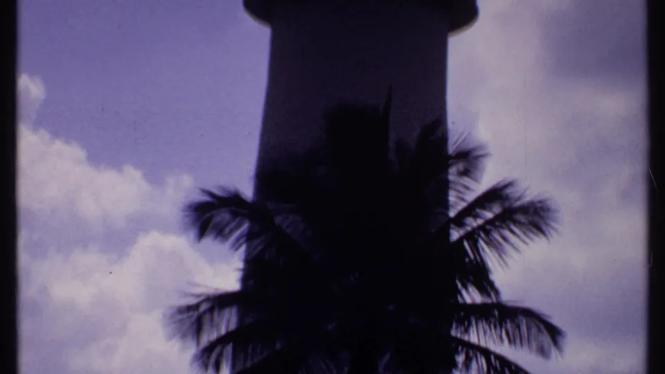 1969 Bahamas 8mm frame with a lighthouse-like tower in silhouette, palm fronds, and purple-tinged clouds.
