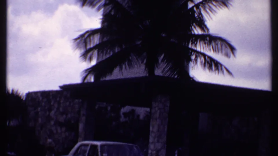 1969 Bahamas 8mm frame showing a palm tree, low stone resort structure, cloudy sky, and a partially visible white car.