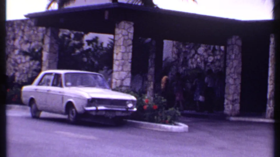 1969 Bahamas 8mm frame of a white sedan beside a stone-columned resort entrance with distant visitors under the canopy.