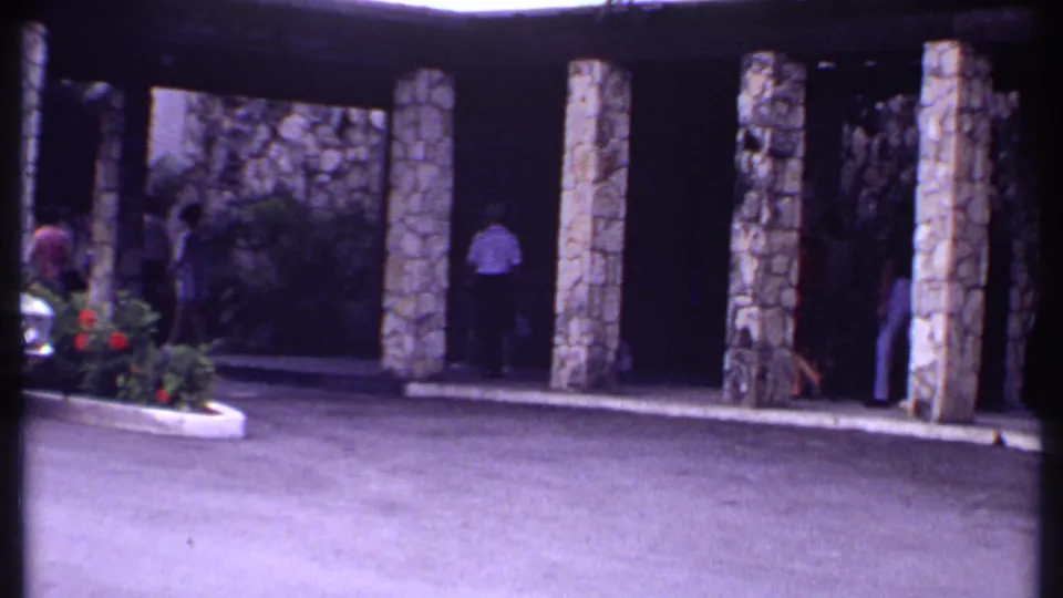 1969 Bahamas home-movie frame with visitors near a stone-pillared covered walkway and a partial parked car at the entrance.