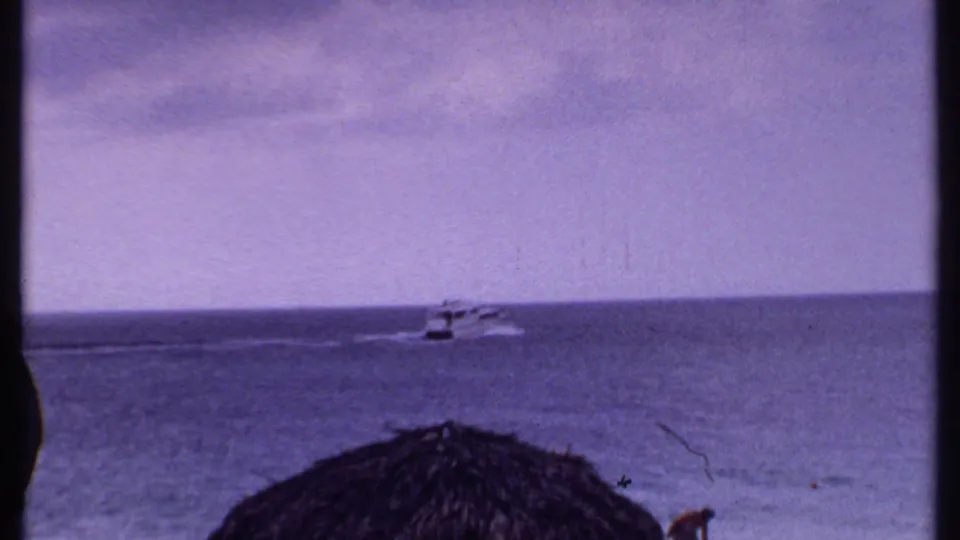 1969 Bahamas home-movie frame showing a small motorboat on open water beneath a hazy horizon, with shore foreground at the bottom.