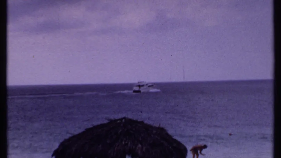 1969 Bahamas home-movie frame of a small motorboat crossing open ocean, with a dark shoreline edge and film gate shadow visible.