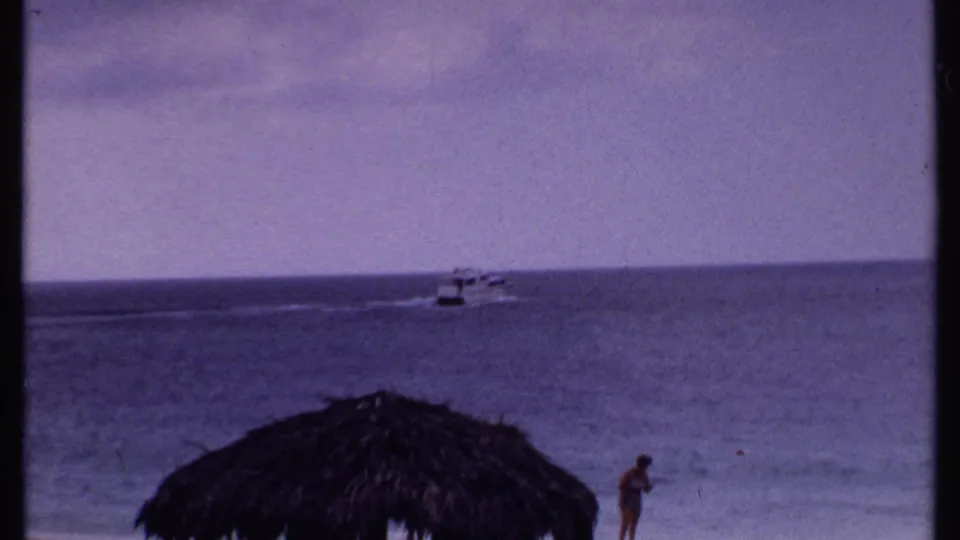 Bahamas home-movie frame from 1969 showing ocean water, a distant boat, a thatched umbrella, and a small figure near the shore.