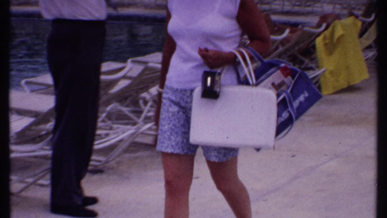 Woman carrying bags near poolside lounge chairs, with another adult partly visible.