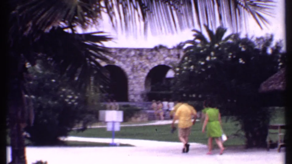 Bahamas 1969 resort frame with two adults walking past palm foliage, white chairs, and a stone archway in the background.