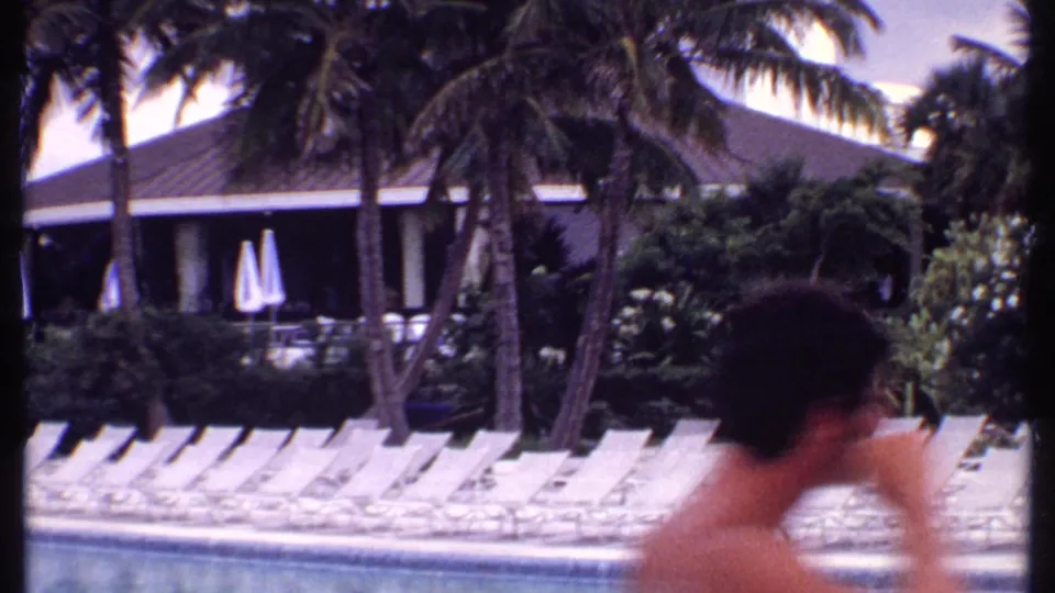 A brief foreground passerby appears at a 1969 Bahamas resort pool, with empty loungers, palms, closed umbrellas, and low buildings behind.
