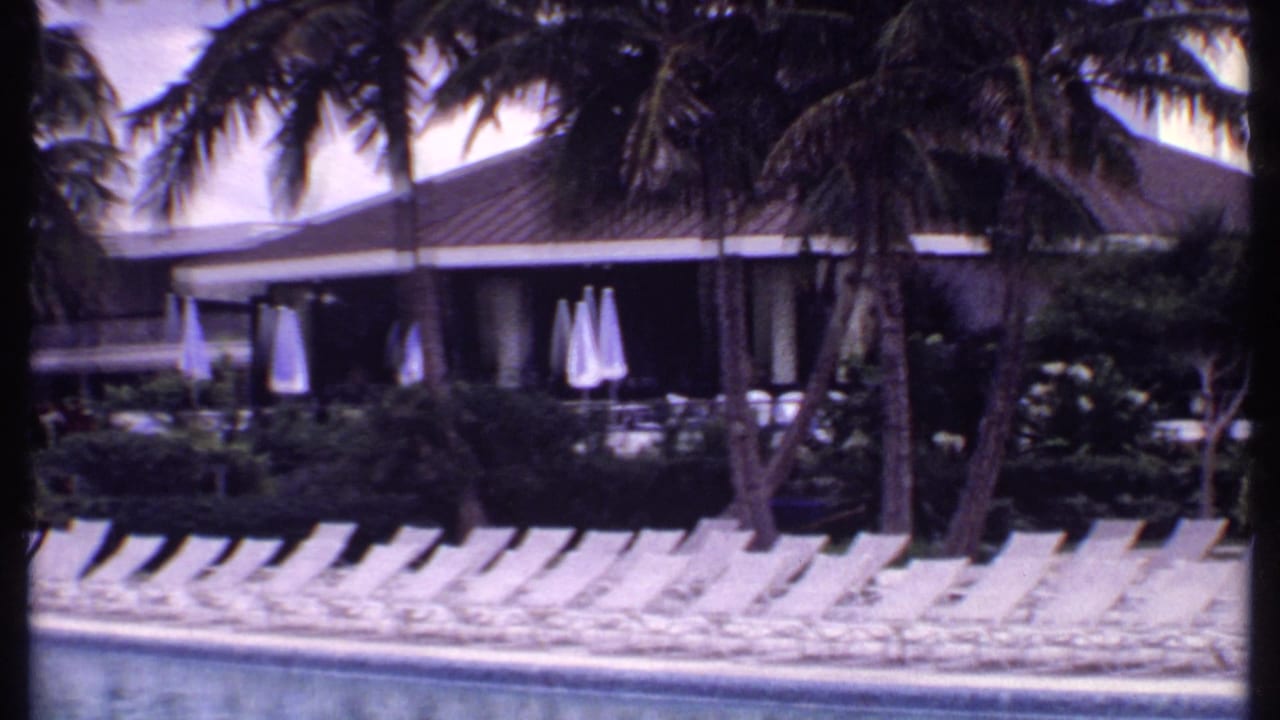 Resort pool deck with white lounge chairs, closed umbrellas, palms, and a low building.