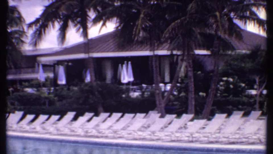 1969 Bahamas resort pool area with empty white loungers, closed umbrellas, palm trees, and a low hotel building beyond the water.