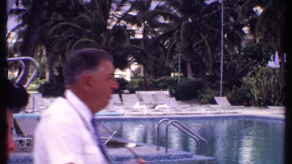 At a 1969 Bahamas resort pool, an adult man in a white shirt and a dark-haired woman pass before loungers, palms, and pool ladders.