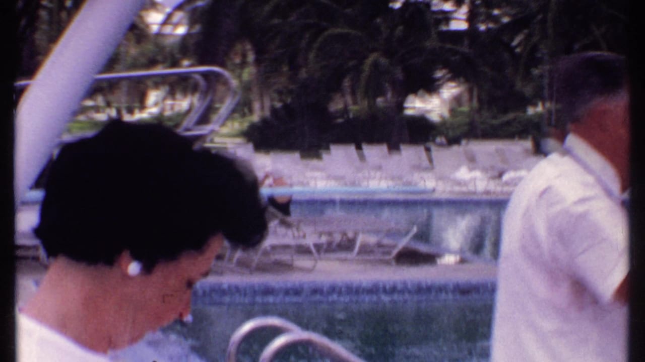 1969 Bahamas home-movie frame showing a woman by a resort swimming pool, with empty loungers, palm trees, and pool railings visible.