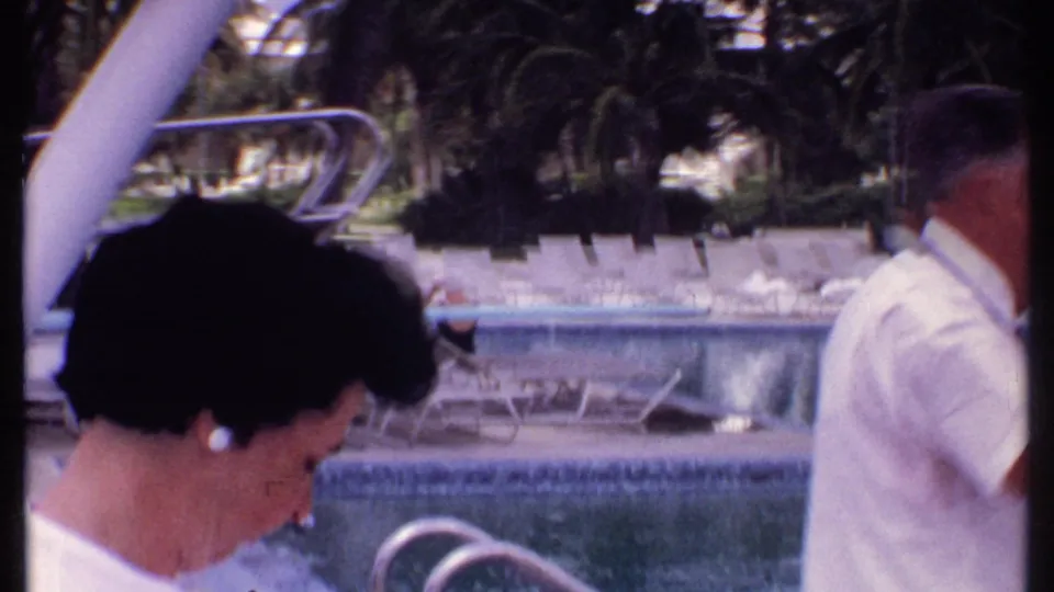 1969 Bahamas home-movie frame showing a woman by a resort swimming pool, with empty loungers, palm trees, and pool railings visible.