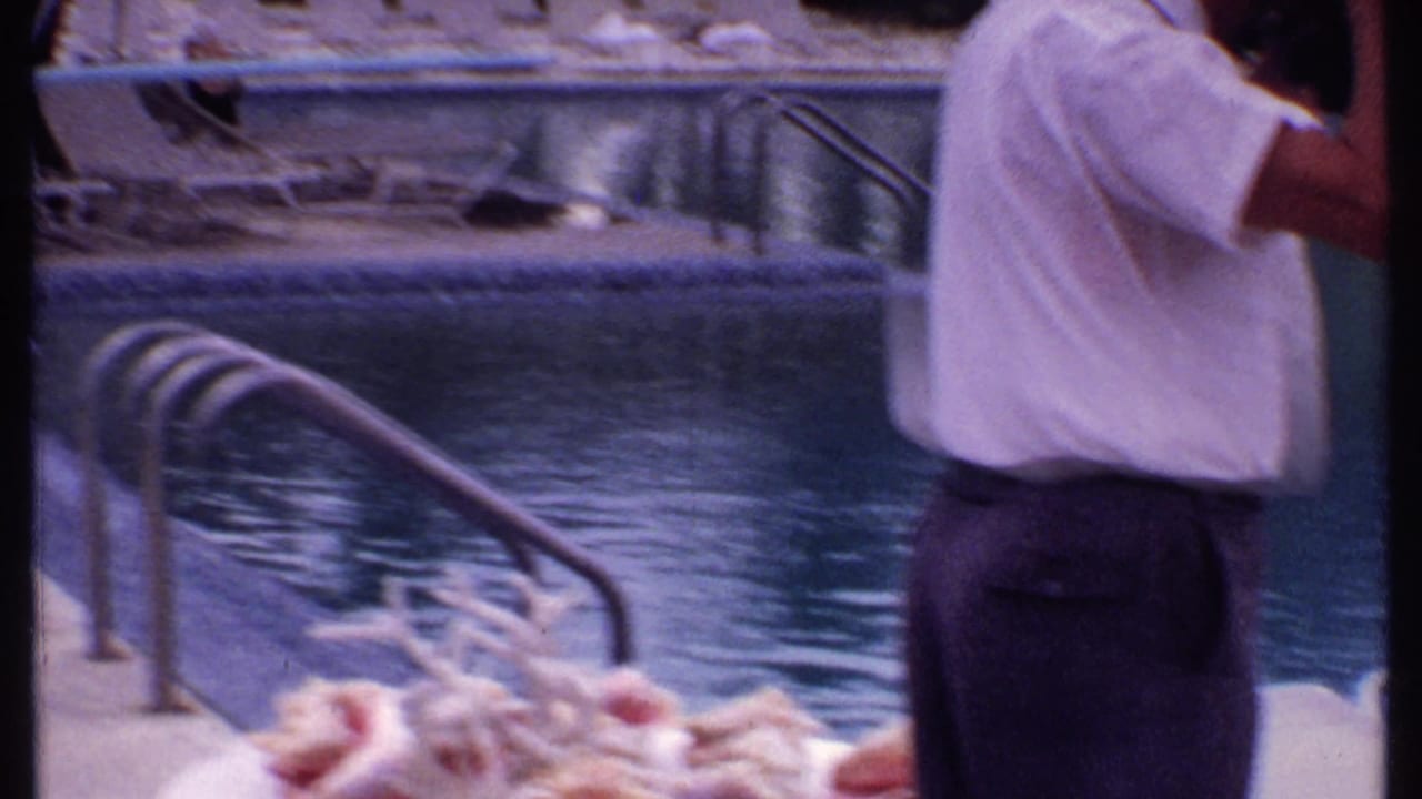Man in a white shirt beside a swimming pool with lounge chairs and palm trees in the background.
