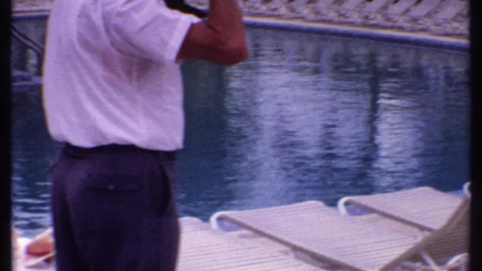 1969 Bahamas home-movie frame showing a man apparently using a camera beside the resort pool, with loungers and blue water visible.