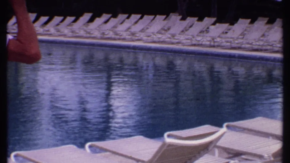 1969 Bahamas resort poolside frame showing blue pool water, rows of empty loungers, and the edge of a man in a white shirt.