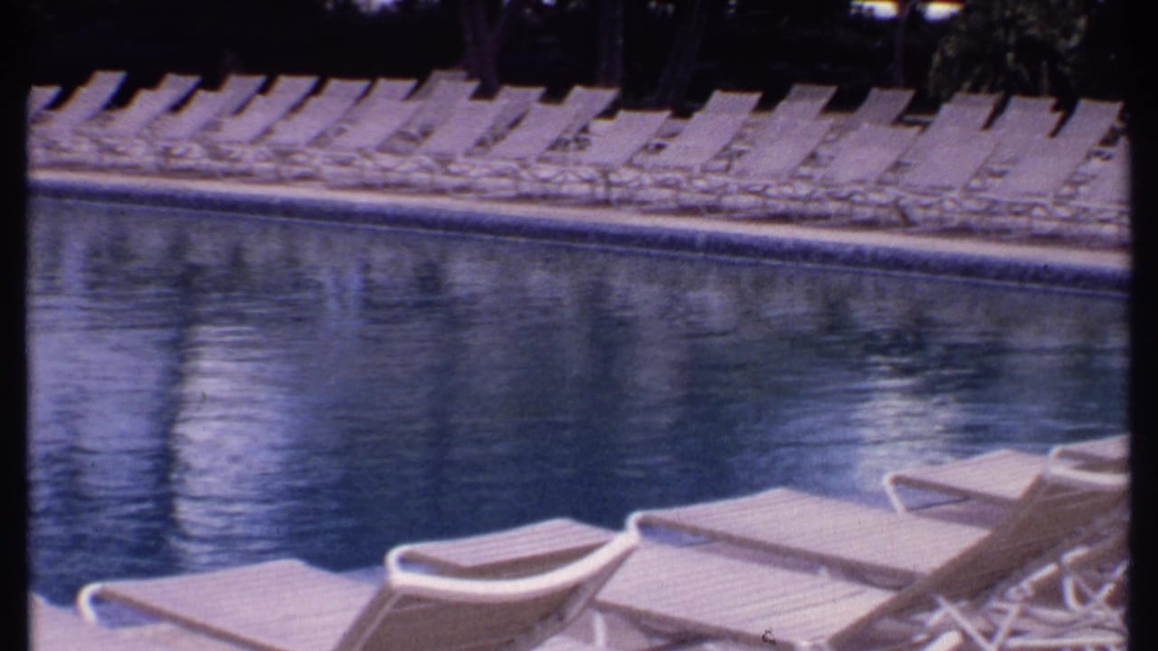 1969 Bahamas home-movie frame showing empty white lounge chairs along a resort pool with shaded edges and water reflections.