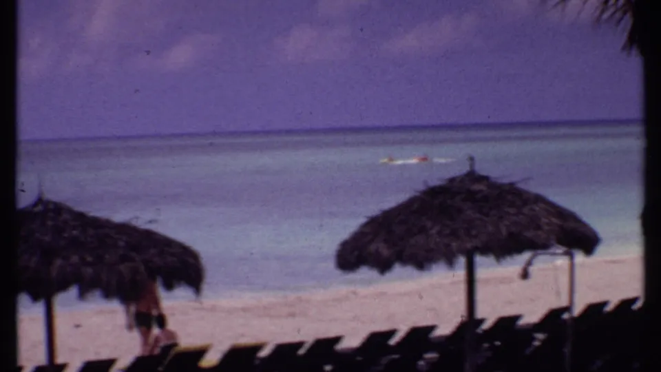 1969 Bahamas resort beach with dark rows of chairs, two thatched umbrellas, and small figures near the shoreline.