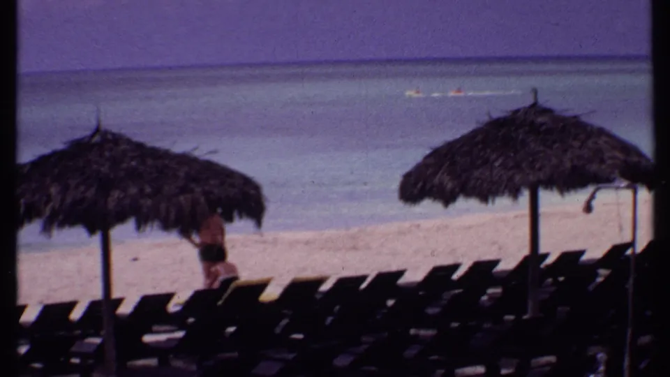 1969 Bahamas home-movie frame showing thatched umbrellas, dark lounge chair rows, calm sea, and a beachgoer near the chairs.