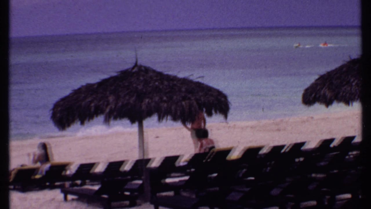 Beach lounge chairs under a thatched umbrella beside calm turquoise water.
