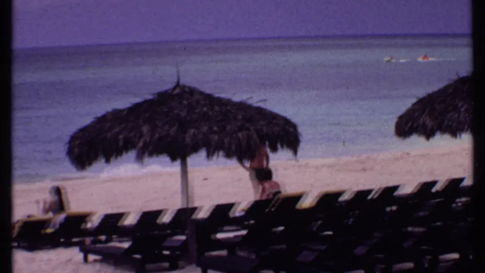 1969 Bahamas beach view with rows of dark lounge chairs, a thatched umbrella, and people relaxing near calm turquoise water.