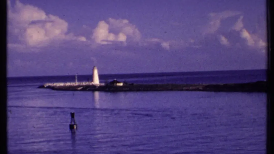 1969 Bahamas harbor view showing a white lighthouse on a rocky jetty under a blue sky with scattered clouds.