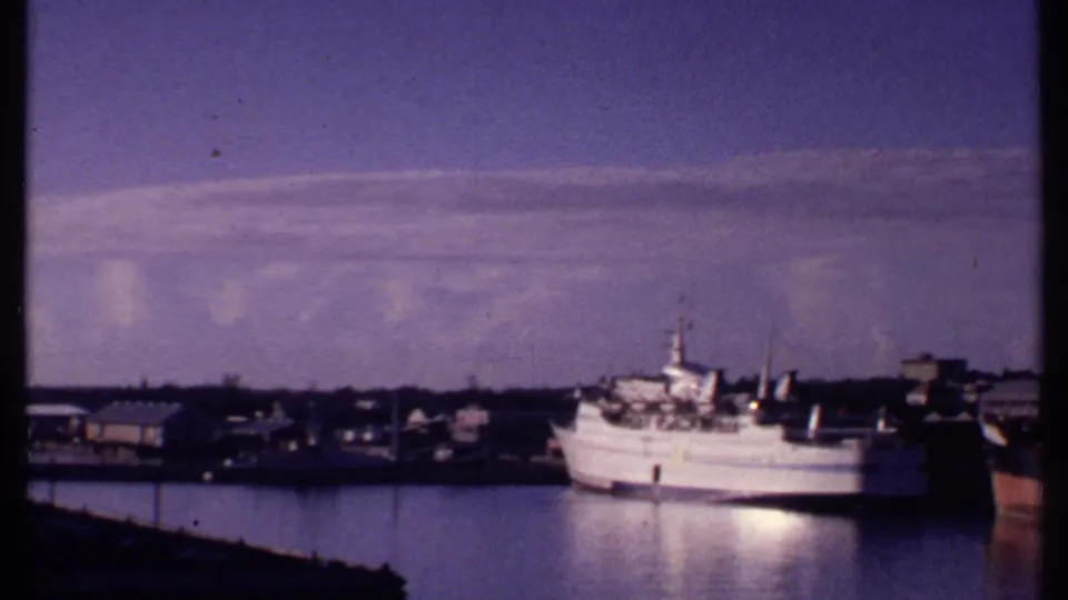 1969 Bahamas harbor frame with a white passenger vessel docked near low waterfront buildings beneath a tinted sky.