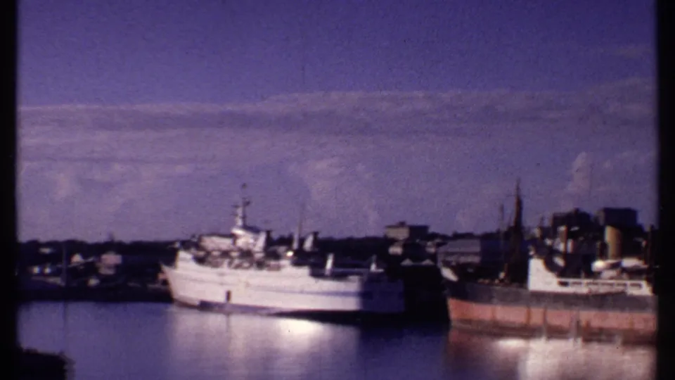 1969 Bahamas harbor frame showing a white vessel beside a rusty cargo ship, cranes, dock buildings, and reflective water.