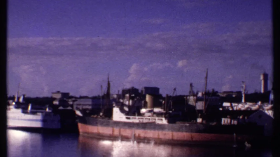 1969 Bahamas harbor frame with a rust-colored cargo ship, white vessel, cranes, dock buildings, and a distant tower.