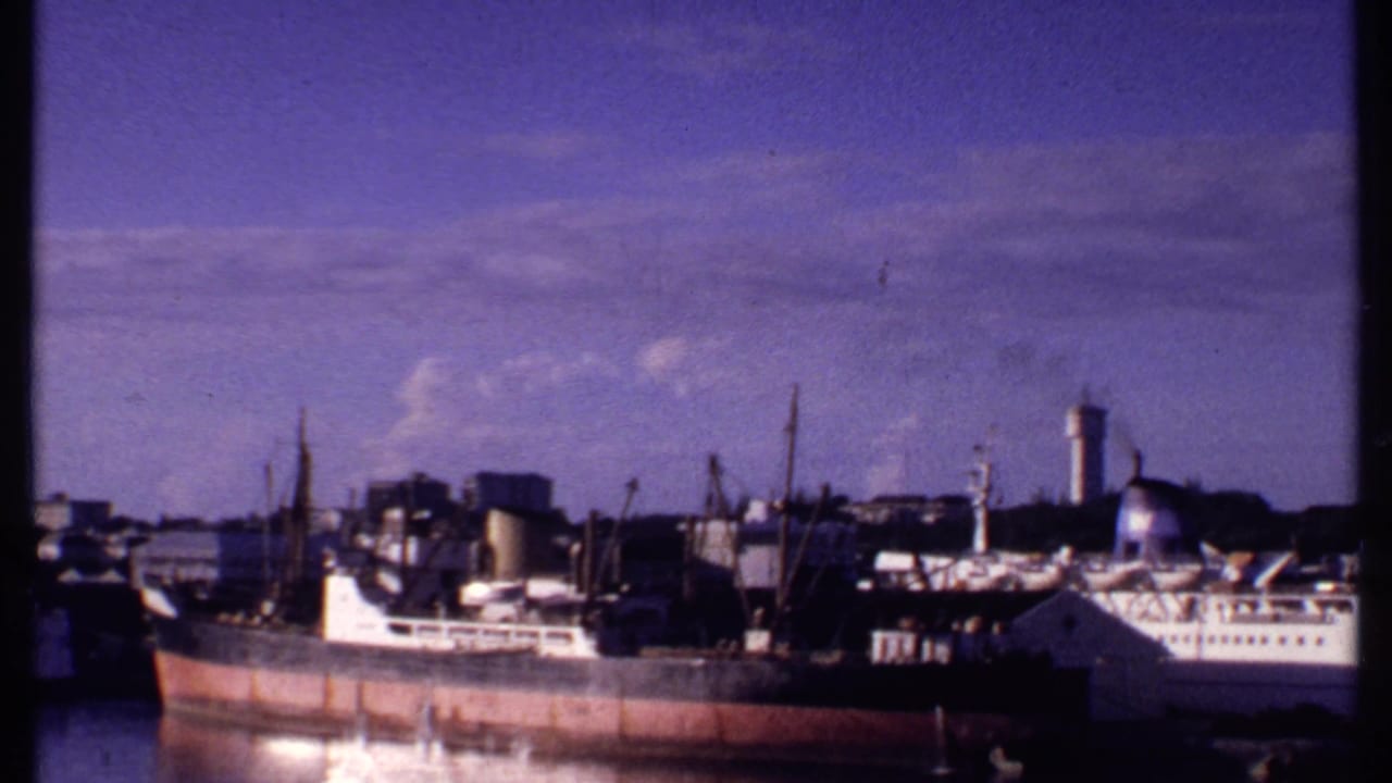 1969 Bahamas harbor view with a white passenger ship near cargo vessels, cranes, industrial buildings, and a water tower.
