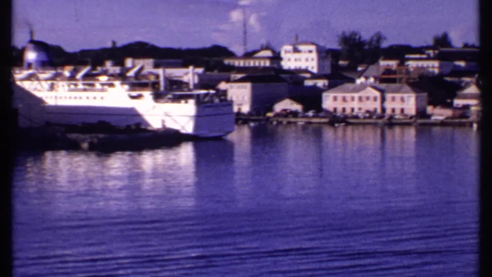 1969 Bahamas port view with a white passenger ship at left, low waterfront buildings, and rippled harbor water in the foreground.