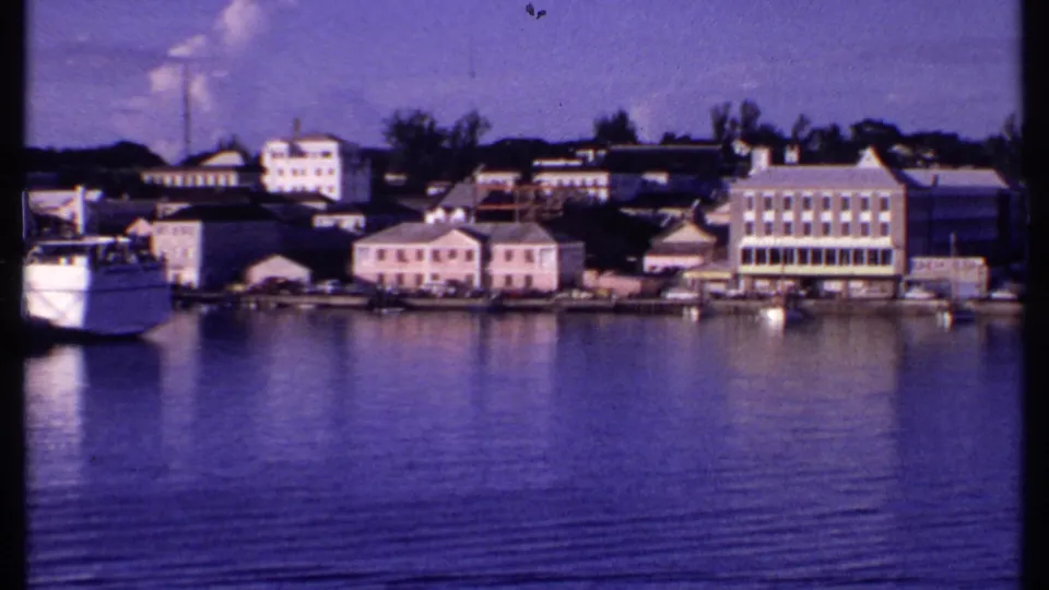 Bahamas waterfront seen across calm water, with a ferry edge, pastel buildings, docks, and trees in the background.