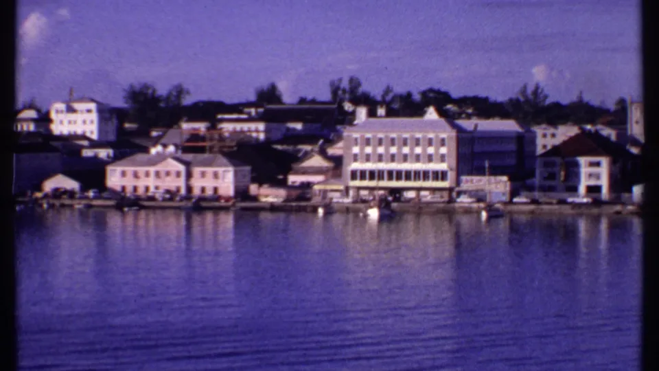 1969 Bahamas harbor shoreline with pale colonial-style buildings, small boats, and reflections on gently rippled water.