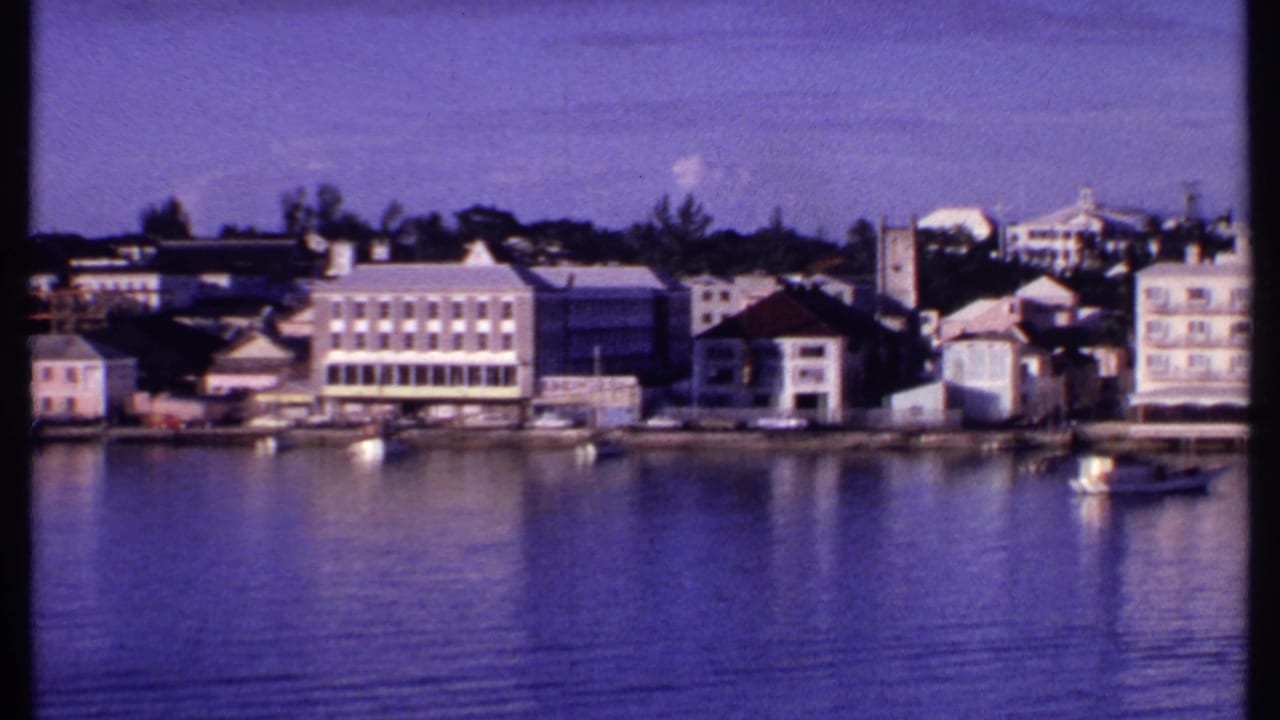 1969 Bahamas 8mm frame showing calm harbor water, pastel buildings along the shoreline, and small boats near the waterfront.