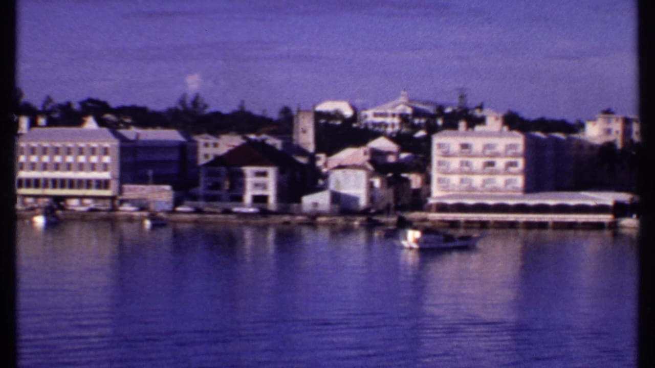 1969 Bahamas harbor view with pastel waterfront buildings, a small boat, and reflections on purple-toned water.