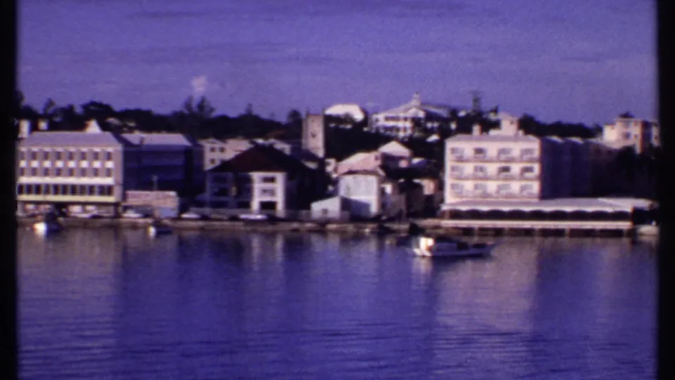 1969 Bahamas harbor view with pastel waterfront buildings, a small boat, and reflections on purple-toned water.