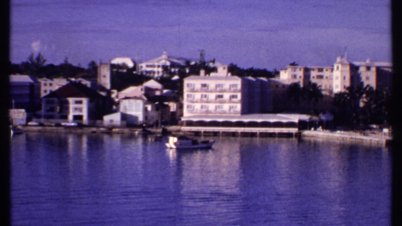 1969 Bahamas 8mm frame with a small boat floating before waterfront buildings, awnings, palms, and still harbor water.