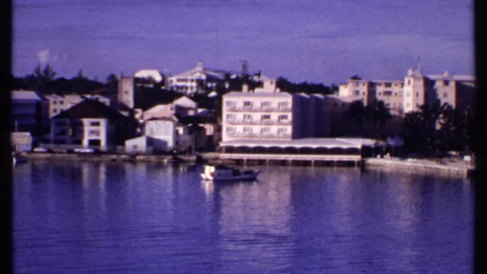1969 Bahamas 8mm frame with a small boat floating before waterfront buildings, awnings, palms, and still harbor water.
