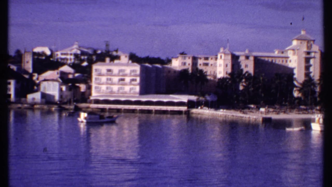 1969 Bahamas waterfront frame showing pastel hotel-like buildings, palms, small boats, and calm harbor water.