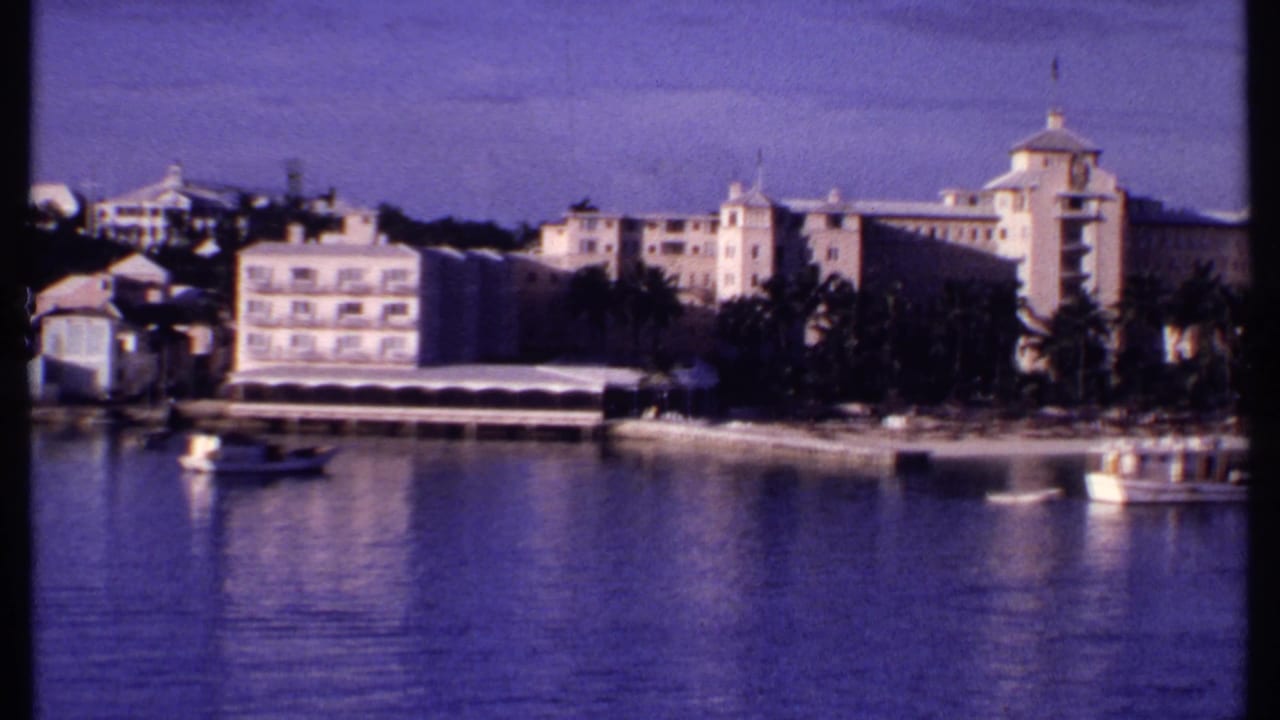 1969 Bahamas home-movie frame showing resort-like waterfront buildings, palms, small boats, and calm harbor water.
