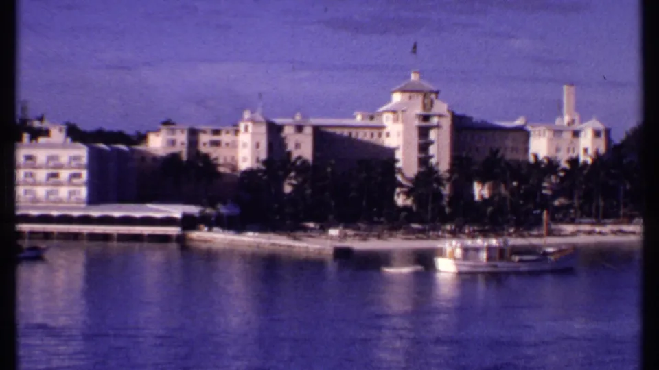 1969 Bahamas waterfront view with pale resort buildings, palm-lined shore, small boats, and reflective blue water.