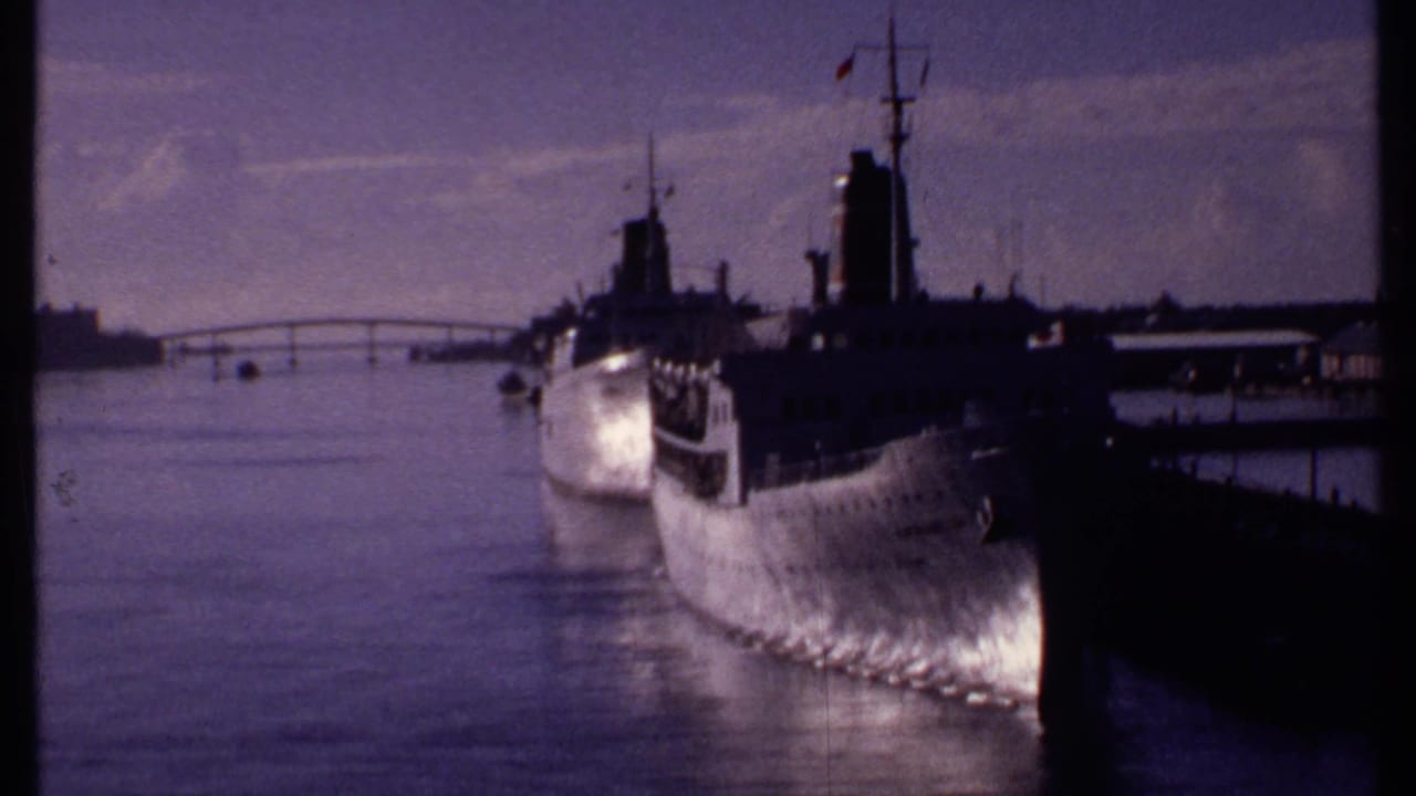 1969 Bahamas home-movie frame with a large docked ship, pier, calm harbor water, and distant low shoreline.