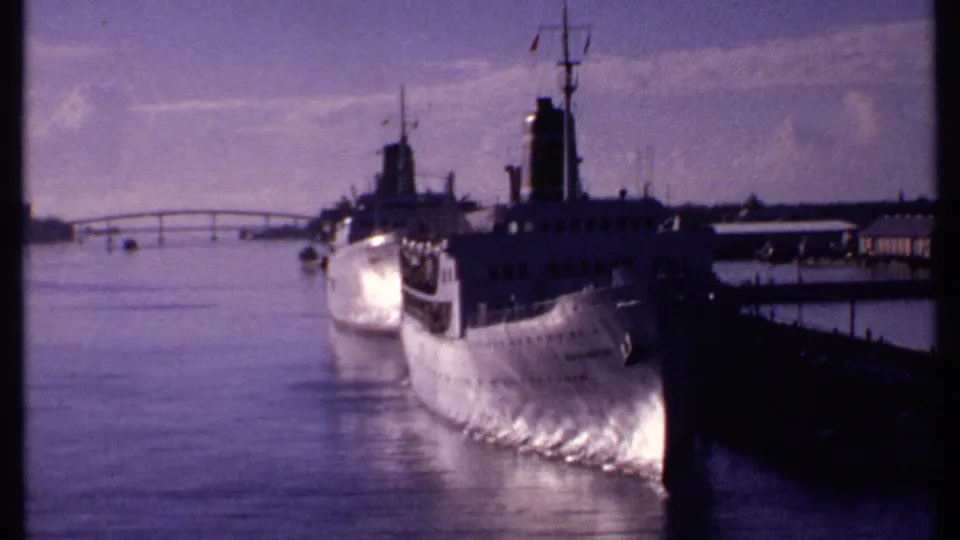 1969 Bahamas 8mm frame showing two large white steamers moored near a dark pier, with a low bridge in the distance.