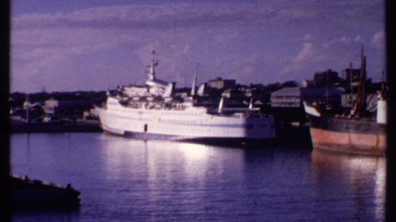 1969 Bahamas waterfront frame showing a white passenger vessel, low-rise harbor buildings, and a darker rust-colored vessel nearby.